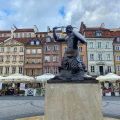 Syrenka Warszawska, jeden z najbardziej rozpoznawalnych pomników w Warszawie, stoi na tle kolorowych kamienic Starego Miasta.