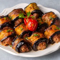 Roladki z bakłażana z mięsem mielonym, polane sosem i posypane natką pietruszki. Obok pieczony pomidorek.