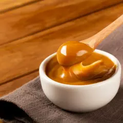 Gęste, karmelowe dulce de leche spływa z drewnianej łyżki do białej miseczki.