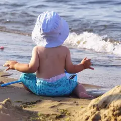 Niemowlak w kapelusiku i kąpielówkach z rekinem siedzi na piasku, patrząc na fale. Idealne miejsce, gdzie nad morze z niemowlakiem.