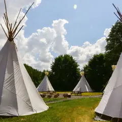 Malowniczy **pole namiotowe** z białymi tipi, drewnianymi ławami i stołami, otoczone zielenią i błękitnym niebem z chmurami.