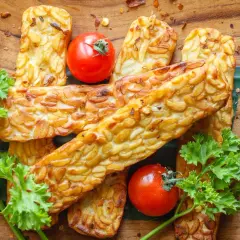Złociste kawałki tempeh, czyli fermentowanej soi, ułożone na drewnianej desce. Obok pomidorki koktajlowe i natka pietruszki.