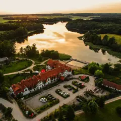 Malowniczy folwark mazurskie legendy nad jeziorem, z dachem krytym czerwoną dachówką, otoczony zielenią.