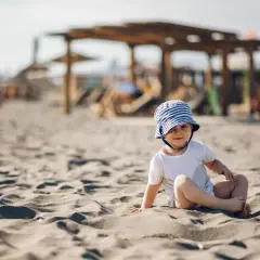 Uroczy maluch w pasiastym kapelusiku siedzi na piaszczystej plaży. Idealne miejsce, gdzie na wakacje z rocznym dzieckiem.
