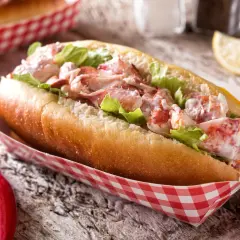 Pyszny lobster roll z kawałkami homara i sałatą w bułce, podany w czerwono-białym papierowym opakowaniu. Obok szczypce do homara.