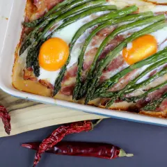 Pyszne szparagi na śniadanie z boczkiem i jajkami zapieczone w naczyniu. Obok leżą suszone papryczki chili.