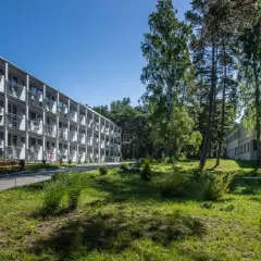 Hotel NAT w Krynicy Morskiej otoczony zielenią, z długim budynkiem o wielu balkonach i drzewami w tle.