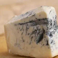 Kawałek kremowego sera gorgonzola z charakterystycznymi niebiesko-zielonymi żyłkami pleśni, leżący na drewnianej desce.