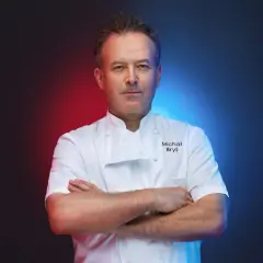 Michał Bryś w białym stroju szefa kuchni, z rękami skrzyżowanymi na piersi, na tle czerwono-niebieskiego gradientu.