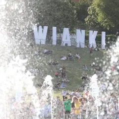 Ludzie siedzą na trawie pod wielkimi literami "Wianki". W tle widać drzewa i fontannę. To radosne wydarzenie, Noc Kupały w Warszawie, przyciągnęło tłumy.