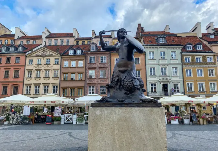 Syrenka Warszawska, jeden z najbardziej rozpoznawalnych pomników w Warszawie, stoi na tle kolorowych kamienic Starego Miasta.