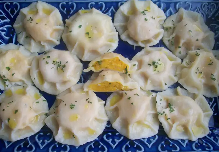 Pyszne pierogi z dynią, polane masłem i posypane szczypiorkiem, ułożone na niebieskiej paterze.