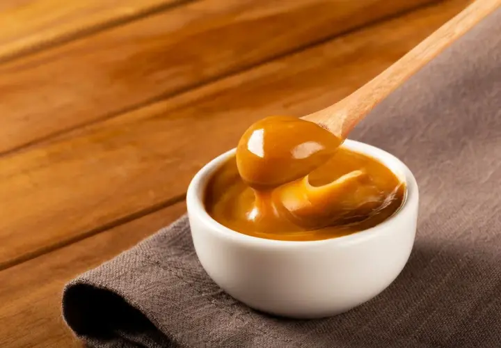Gęste, karmelowe dulce de leche spływa z drewnianej łyżki do białej miseczki.
