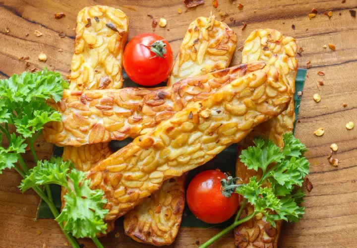 Złociste kawałki tempeh, czyli fermentowanej soi, ułożone na drewnianej desce. Obok pomidorki koktajlowe i natka pietruszki.