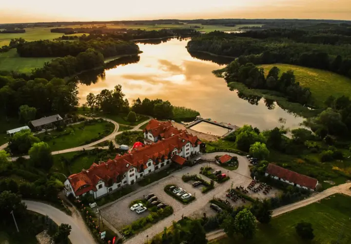 Malowniczy folwark mazurskie legendy nad jeziorem, z dachem krytym czerwoną dachówką, otoczony zielenią.