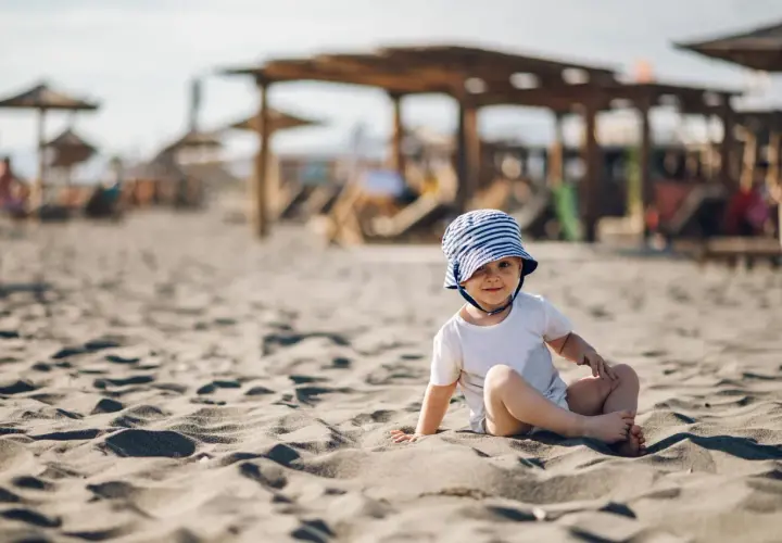Uroczy maluch w pasiastym kapelusiku siedzi na piaszczystej plaży. Idealne miejsce, gdzie na wakacje z rocznym dzieckiem.