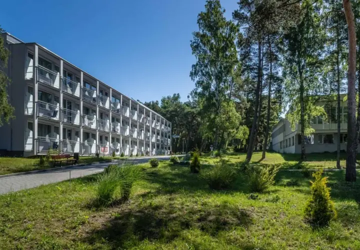 Hotel NAT w Krynicy Morskiej otoczony zielenią, z długim budynkiem o wielu balkonach i drzewami w tle.