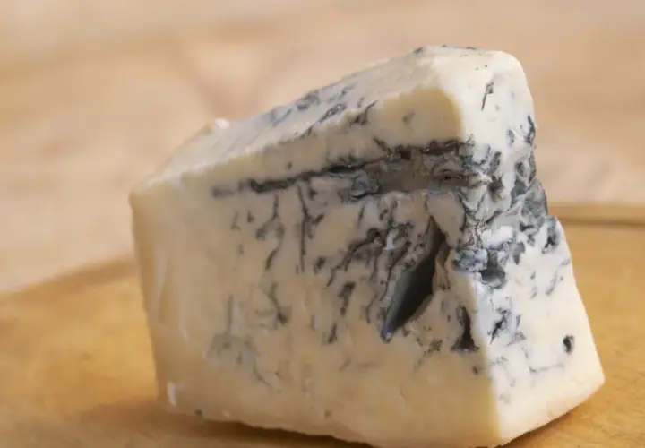 Kawałek kremowego sera gorgonzola z charakterystycznymi niebiesko-zielonymi żyłkami pleśni, leżący na drewnianej desce.