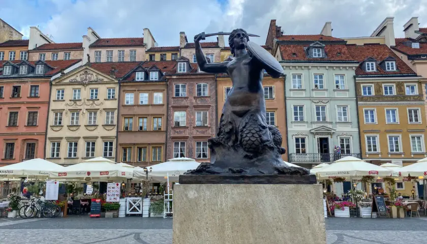 Syrenka Warszawska, jeden z najbardziej rozpoznawalnych pomników w Warszawie, stoi na tle kolorowych kamienic Starego Miasta.