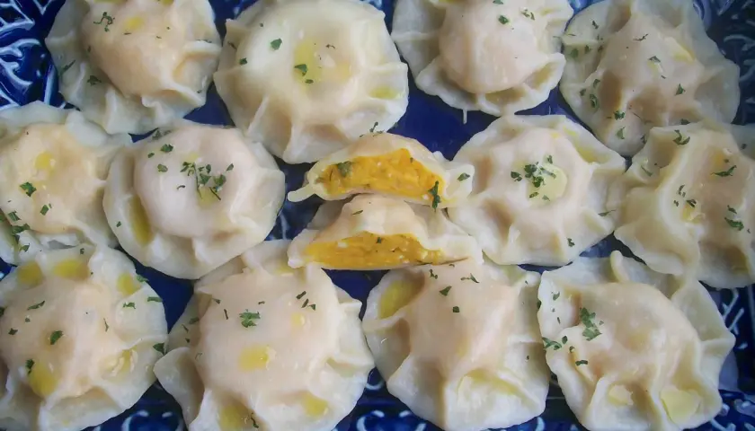 Pyszne pierogi z dynią, polane masłem i posypane szczypiorkiem, ułożone na niebieskiej paterze.