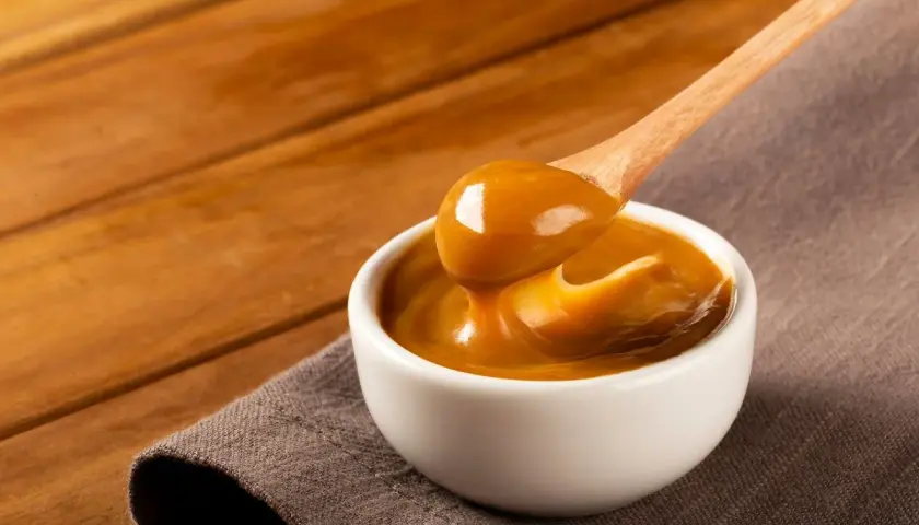 Gęste, karmelowe dulce de leche spływa z drewnianej łyżki do białej miseczki.
