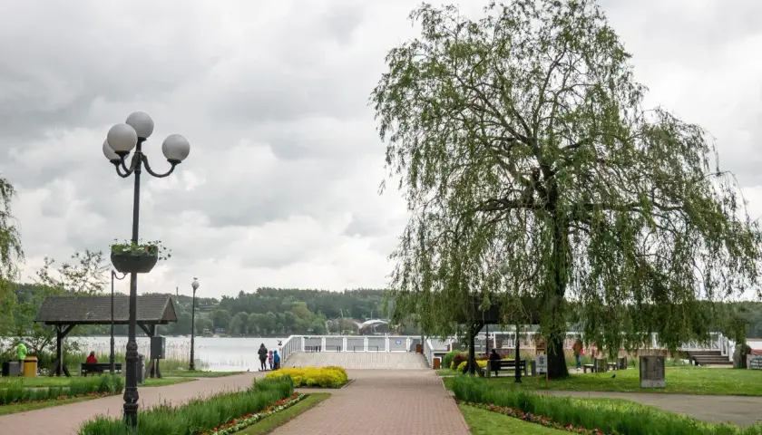 Spacer w Mrągowie nad jeziorem, z wierzbą płaczącą i kwitnącymi rabatami. Idealne miejsce na relaks i podziwianie mrągowskich atrakcji.