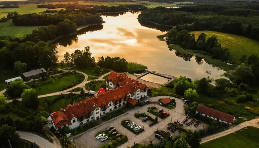 Malowniczy folwark mazurskie legendy nad jeziorem, z dachem krytym czerwoną dachówką, otoczony zielenią.