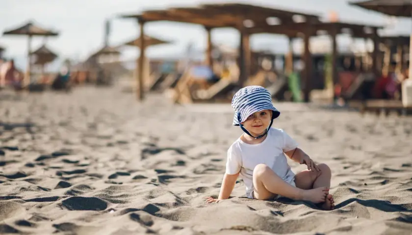 Uroczy maluch w pasiastym kapelusiku siedzi na piaszczystej plaży. Idealne miejsce, gdzie na wakacje z rocznym dzieckiem.