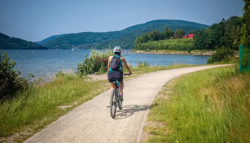 Kobieta na rowerze przemierza malownicze trasy rowerowe nad jeziorem, otoczona zielenią i górami.