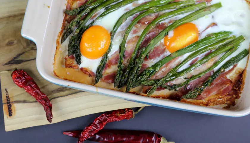 Pyszne szparagi na śniadanie z boczkiem i jajkami zapieczone w naczyniu. Obok leżą suszone papryczki chili.
