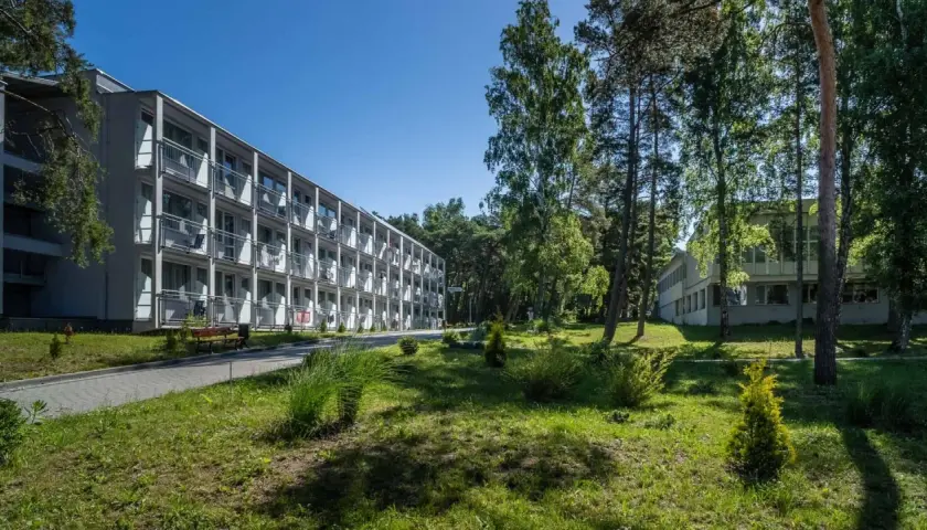 Hotel NAT w Krynicy Morskiej otoczony zielenią, z długim budynkiem o wielu balkonach i drzewami w tle.