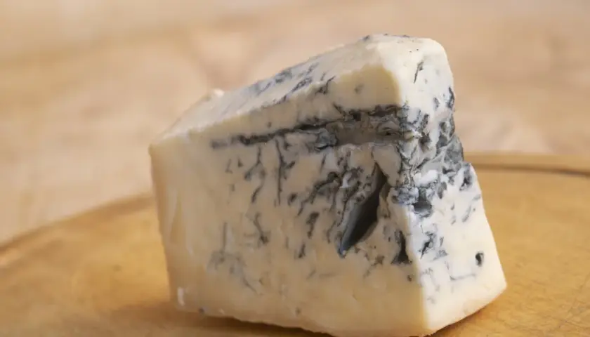 Kawałek kremowego sera gorgonzola z charakterystycznymi niebiesko-zielonymi żyłkami pleśni, leżący na drewnianej desce.