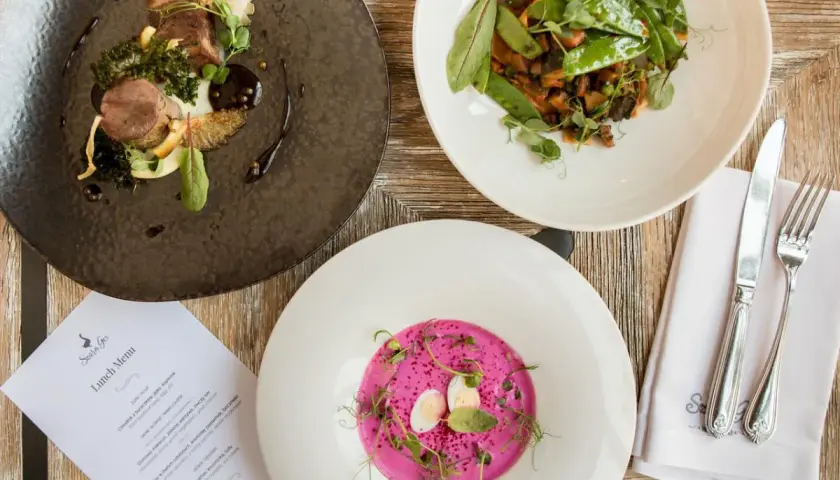 Eleganckie dania na drewnianym stole: mięso z dodatkami, warzywa z groszkiem i różowy chłodnik. Menu "Szara Gęś" zapowiada ucztę.