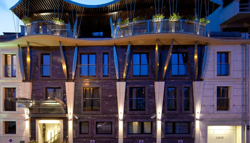 Nowoczesny hotel niebieski z falistym dachem i kamienną fasadą. Wejście oświetlone, obok garaż.