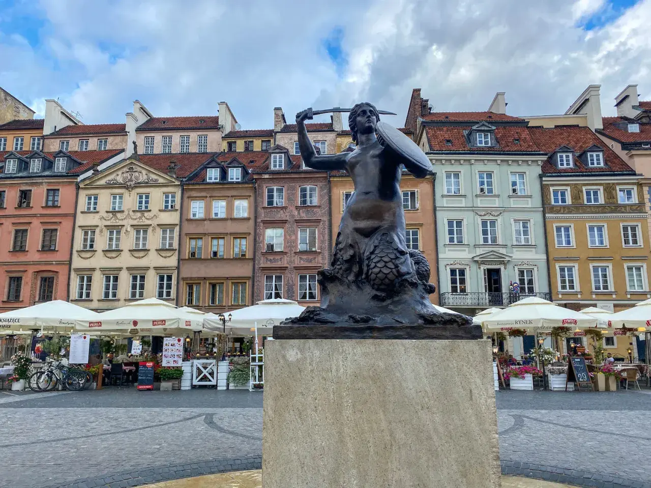 Syrenka Warszawska, jeden z najbardziej rozpoznawalnych pomników w Warszawie, stoi na tle kolorowych kamienic Starego Miasta.