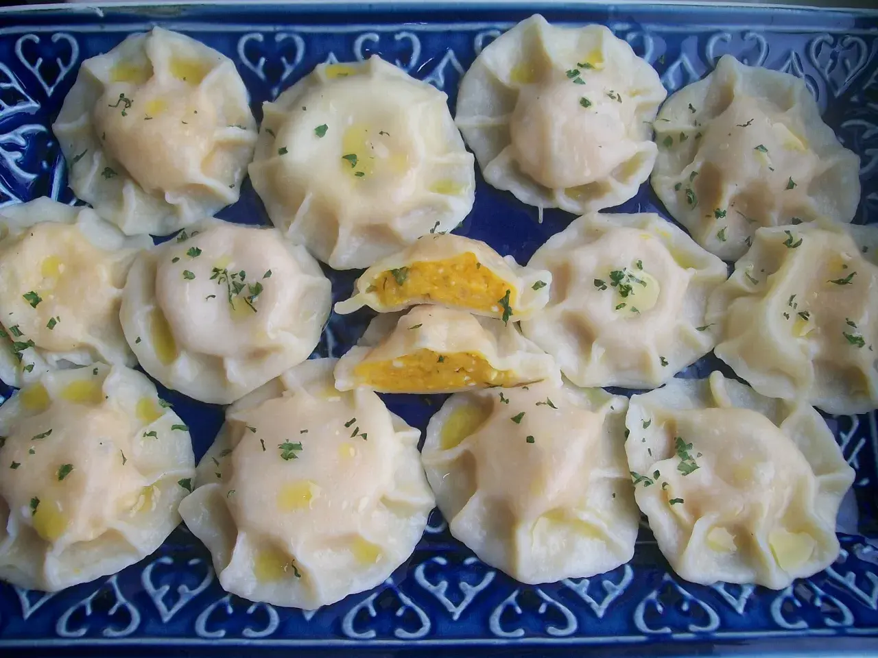 Pyszne pierogi z dynią, polane masłem i posypane szczypiorkiem, ułożone na niebieskiej paterze.