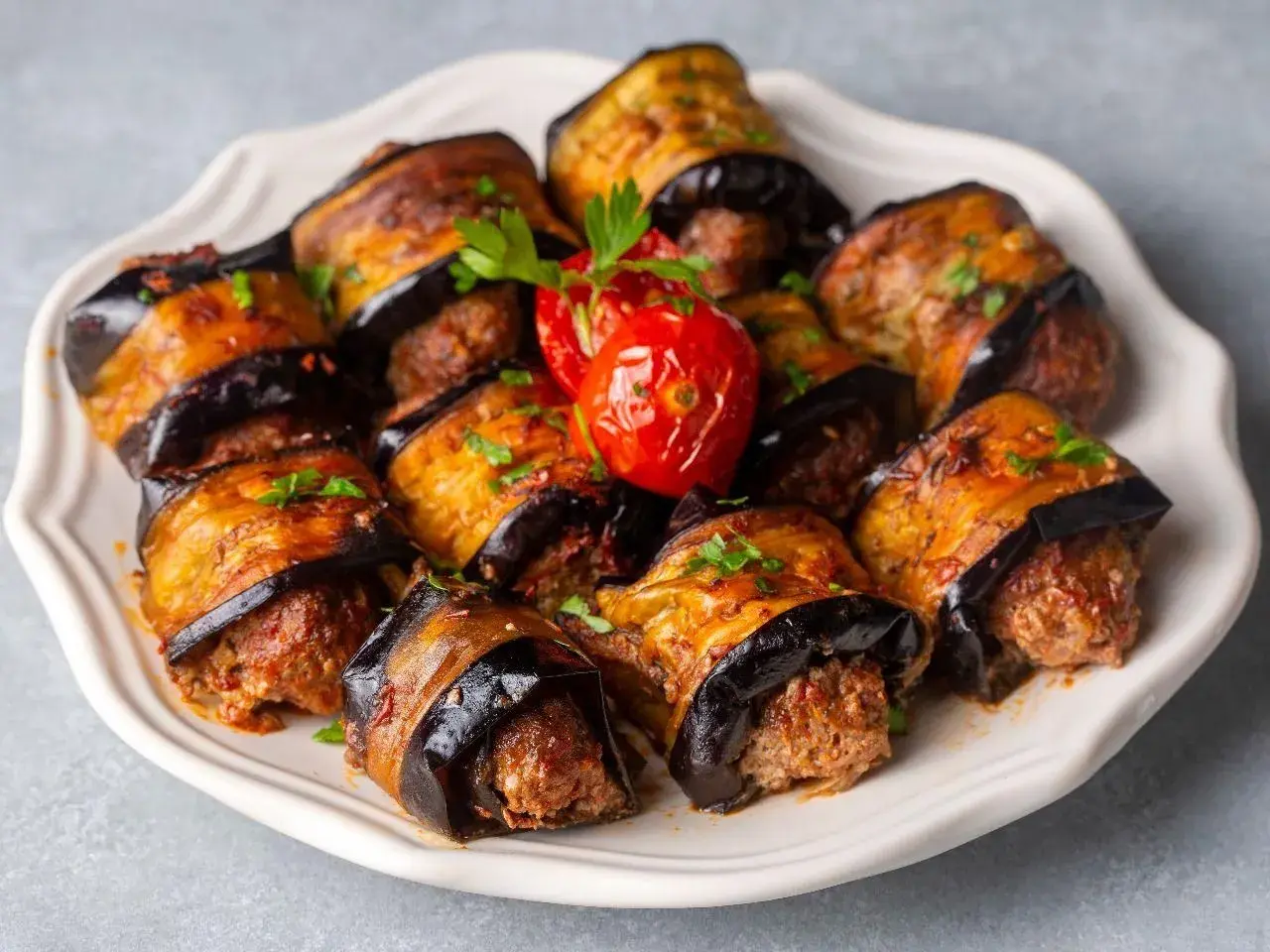 Roladki z bakłażana z mięsem mielonym, polane sosem i posypane natką pietruszki. Obok pieczony pomidorek.