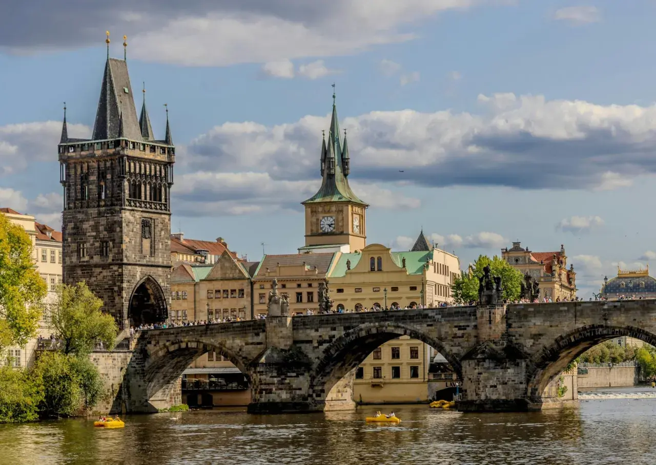 Most piękne stolice Europy, Most Karola w Pradze, z wieżami i zegarem, na tle błękitnego nieba z chmurami.
