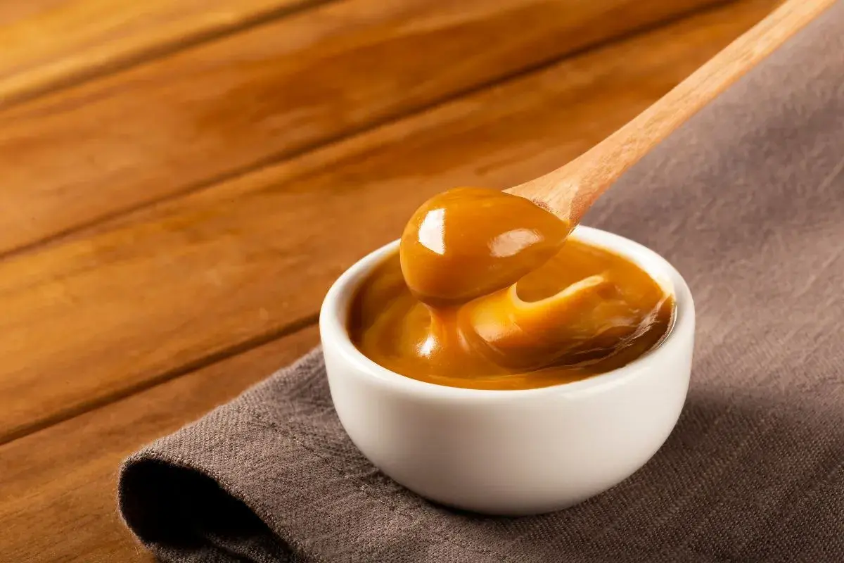 Gęste, karmelowe dulce de leche spływa z drewnianej łyżki do białej miseczki.