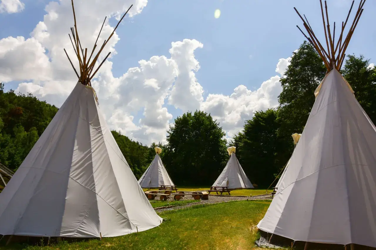 Malowniczy **pole namiotowe** z białymi tipi, drewnianymi ławami i stołami, otoczone zielenią i błękitnym niebem z chmurami.
