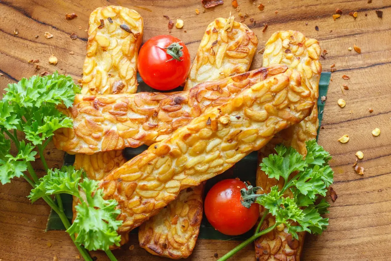 Złociste kawałki tempeh, czyli fermentowanej soi, ułożone na drewnianej desce. Obok pomidorki koktajlowe i natka pietruszki.