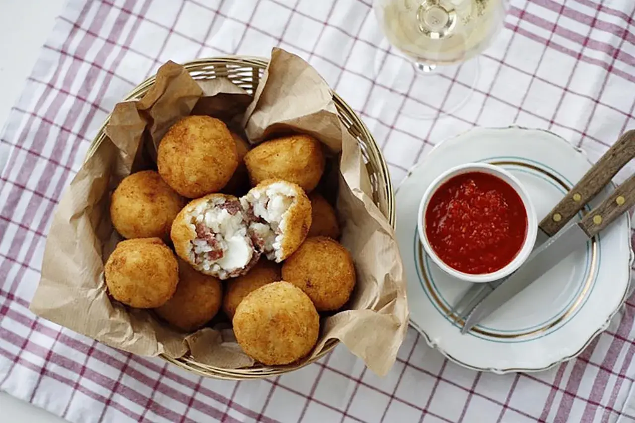 Koszyk pełen złotych arancini, jedno przekrojone ukazuje ryż i ser. Obok miseczka z sosem pomidorowym. Idealne na przekąskę, gdy masz ochotę na arancini przepis.
