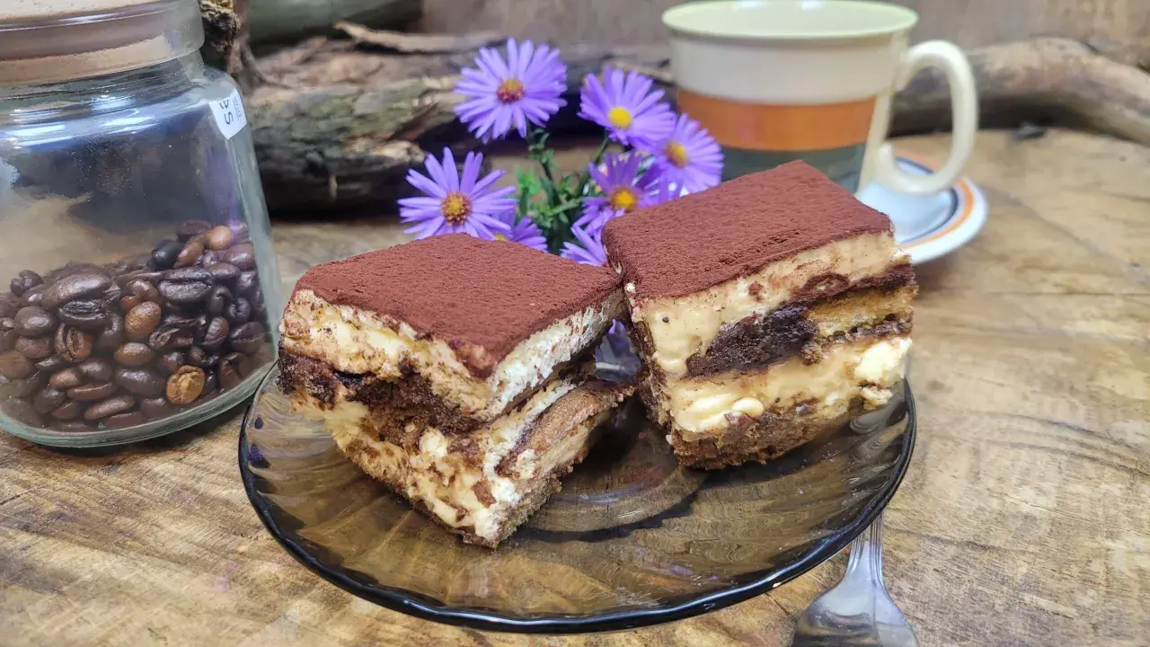 Dwa kawałki tiramisu, idealne ciasto do kawy, obok słoika z ziarnami kawy i fioletowych kwiatów.