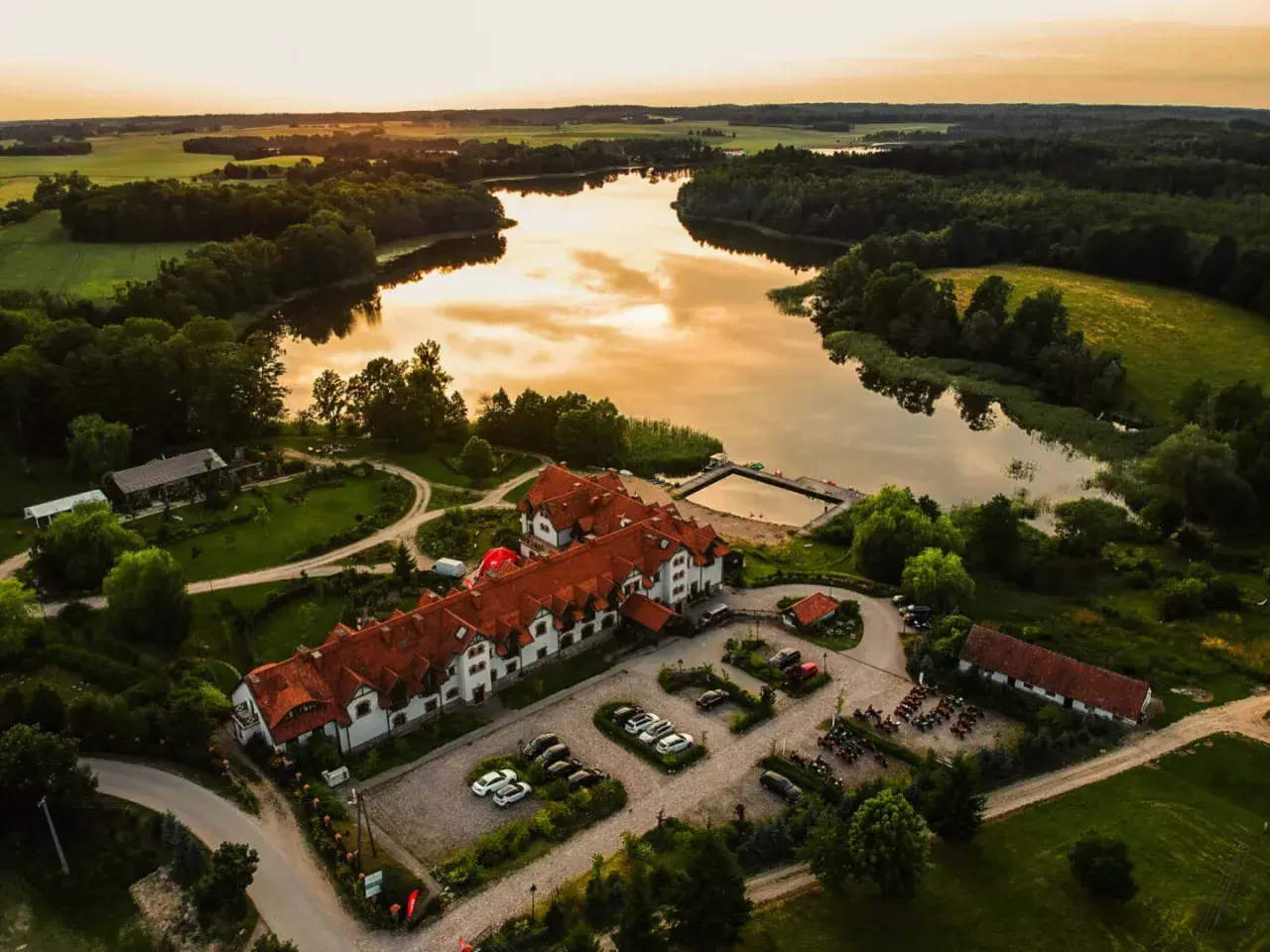 Malowniczy folwark mazurskie legendy nad jeziorem, z dachem krytym czerwoną dachówką, otoczony zielenią.