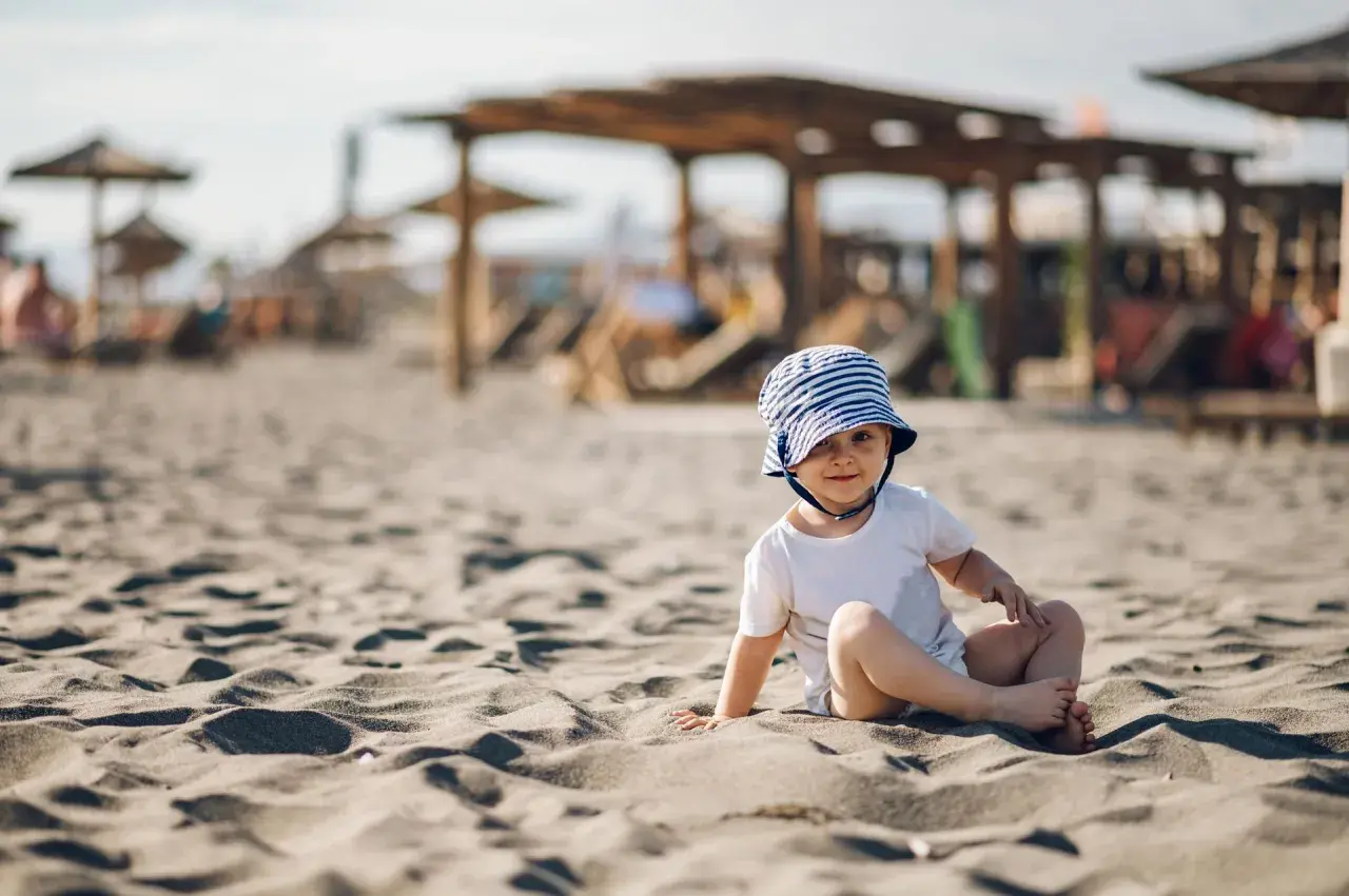 Uroczy maluch w pasiastym kapelusiku siedzi na piaszczystej plaży. Idealne miejsce, gdzie na wakacje z rocznym dzieckiem.