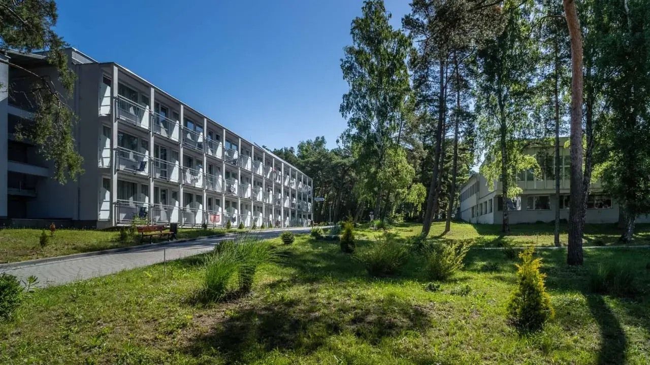 Hotel NAT w Krynicy Morskiej otoczony zielenią, z długim budynkiem o wielu balkonach i drzewami w tle.