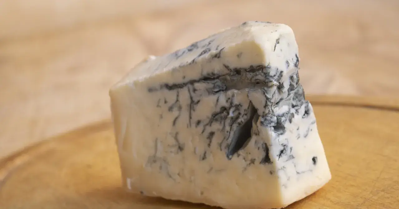 Kawałek kremowego sera gorgonzola z charakterystycznymi niebiesko-zielonymi żyłkami pleśni, leżący na drewnianej desce.