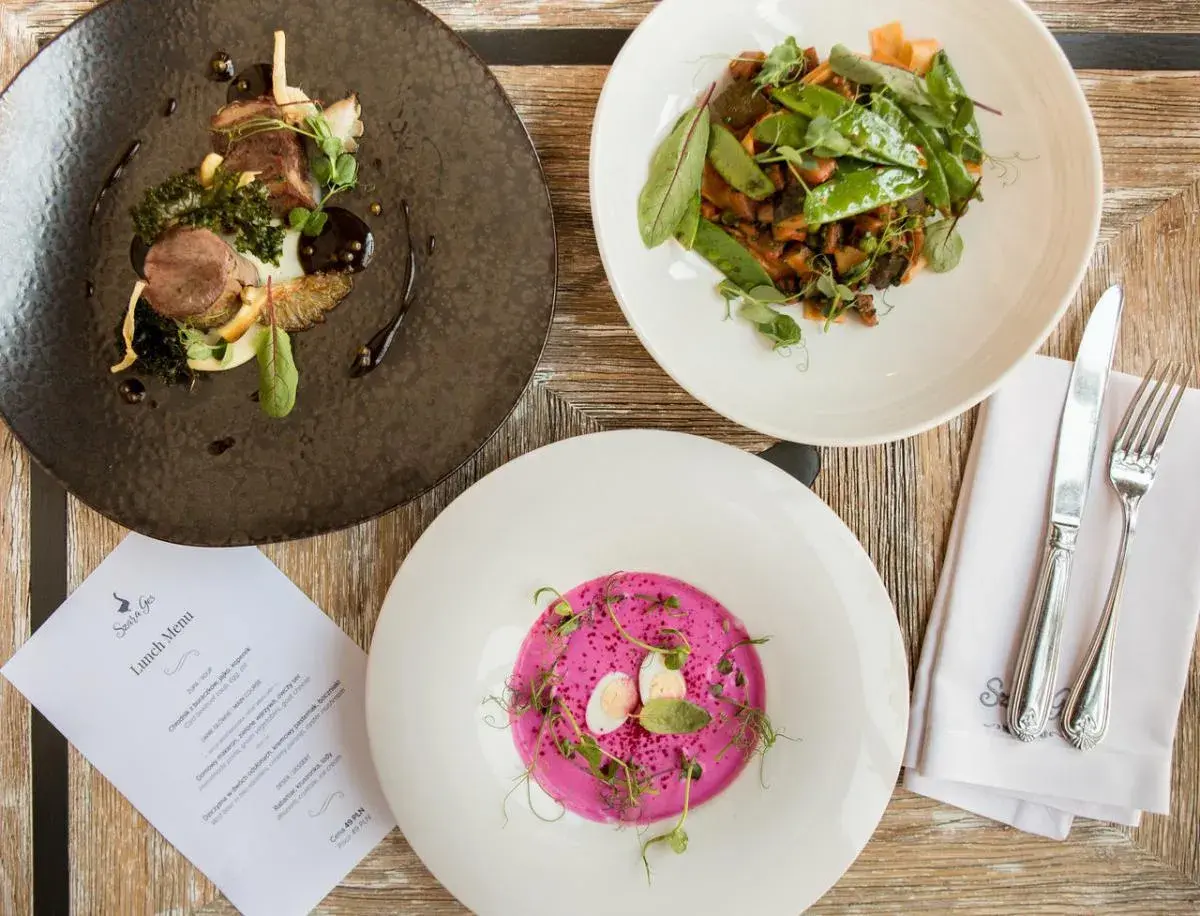 Eleganckie dania na drewnianym stole: mięso z dodatkami, warzywa z groszkiem i różowy chłodnik. Menu "Szara Gęś" zapowiada ucztę.