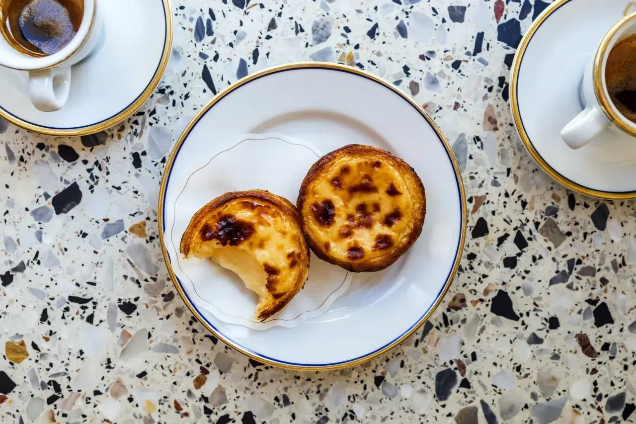 Dwa pyszne pastel de nata na talerzyku, obok filiżanki kawy. Idealny deser na popołudnie.
