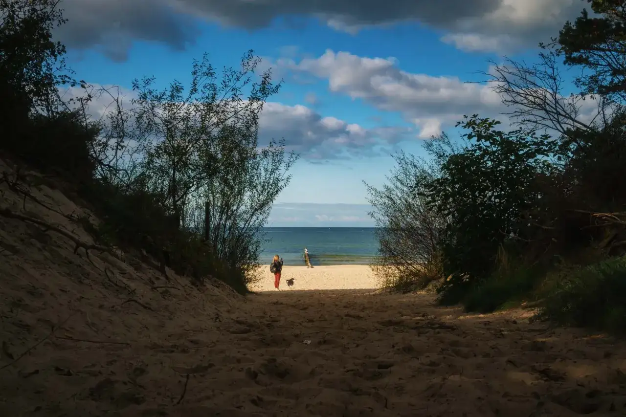 Ścieżka przez wydmy prowadzi do plaży w Pinea Resort Pobierowo. W oddali widać morze i dwie postacie z psem.